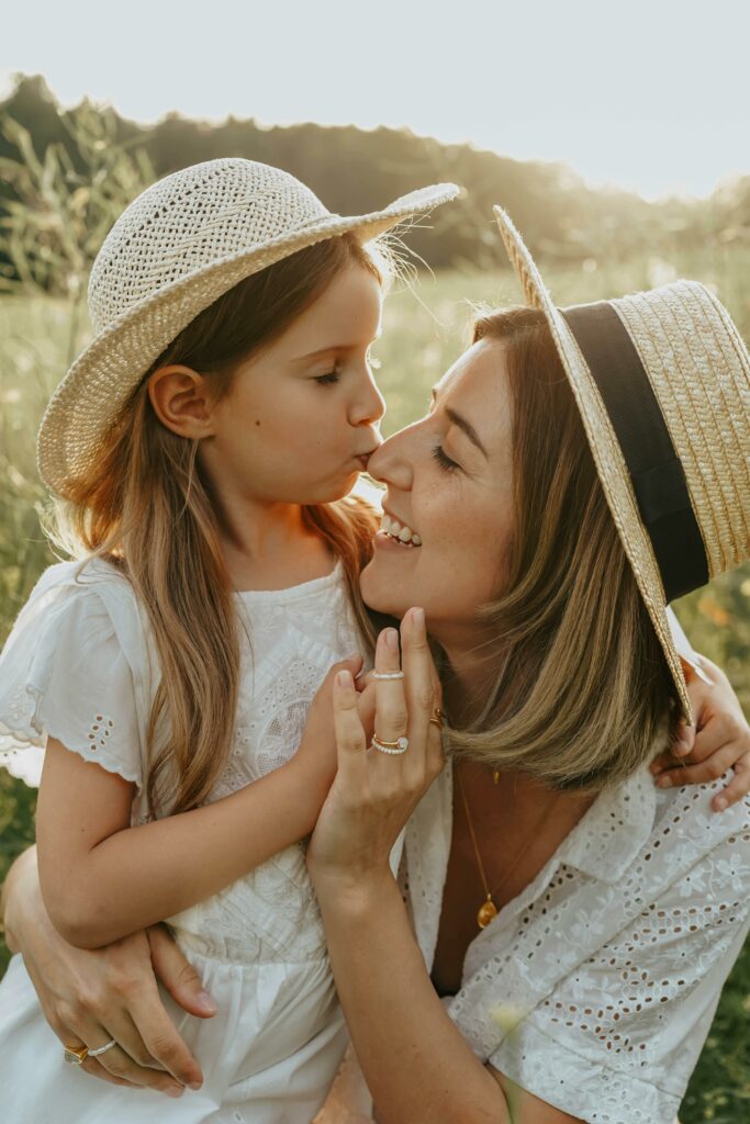 Heartwarming moment of a mother and daughter sharing a kiss in a sunlit field, wearing matching sun hats.